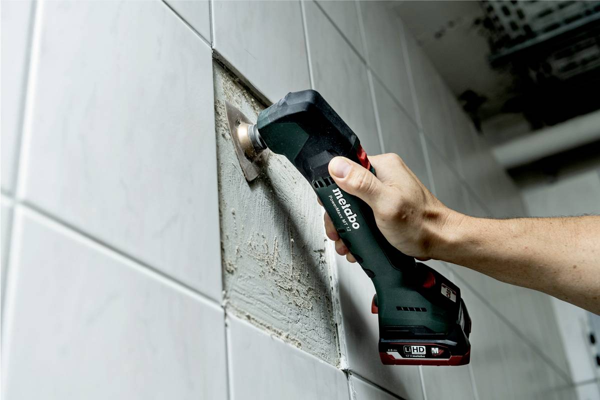 Eine Person entfernt eine Fliese mit einem elektrischen Werkzeug von einer Badezimmerwand.