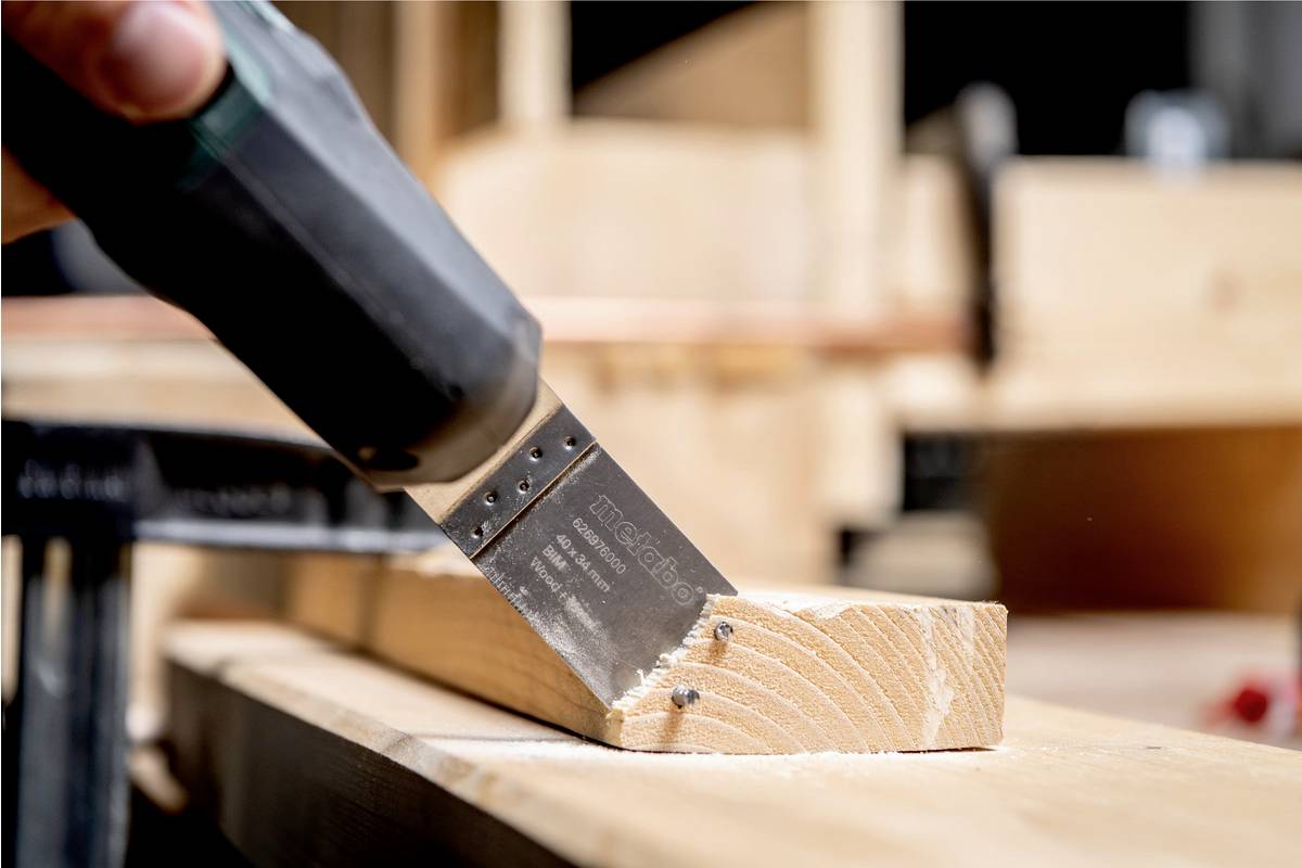 Eine Hand hält eine elektrische Säge und schneidet durch ein Stück Holz. Hintergrund zeigt unscharfe Werkstattumgebung.