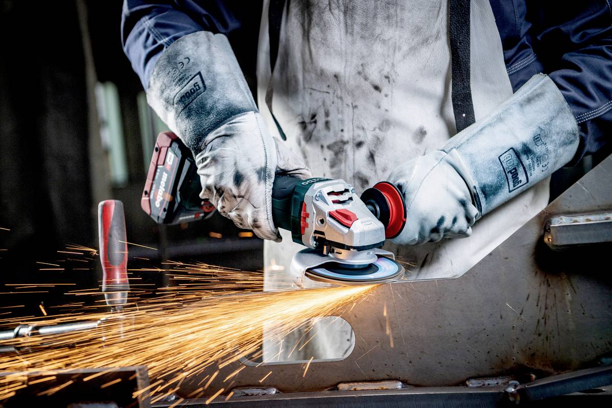 Ein Arbeiter schleift Metall mit einem Winkelschleifer, Funken sprühen. Er trägt Schutzhandschuhe und eine Schürze.