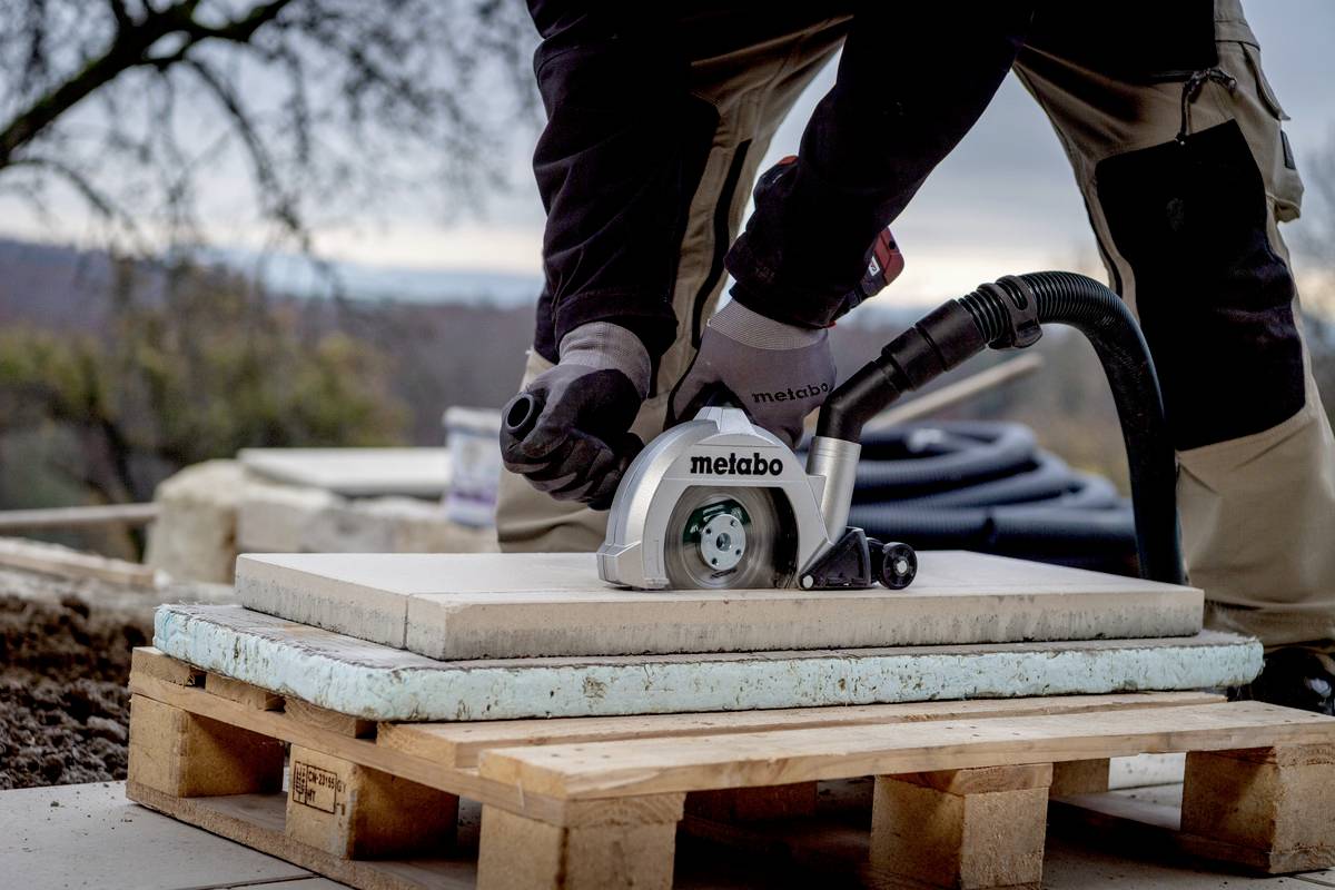 Eine Person schneidet mit einer elektrischen Säge eine Fliese auf einer Baustelle. Die Fliese liegt auf einer Palette im Freien.