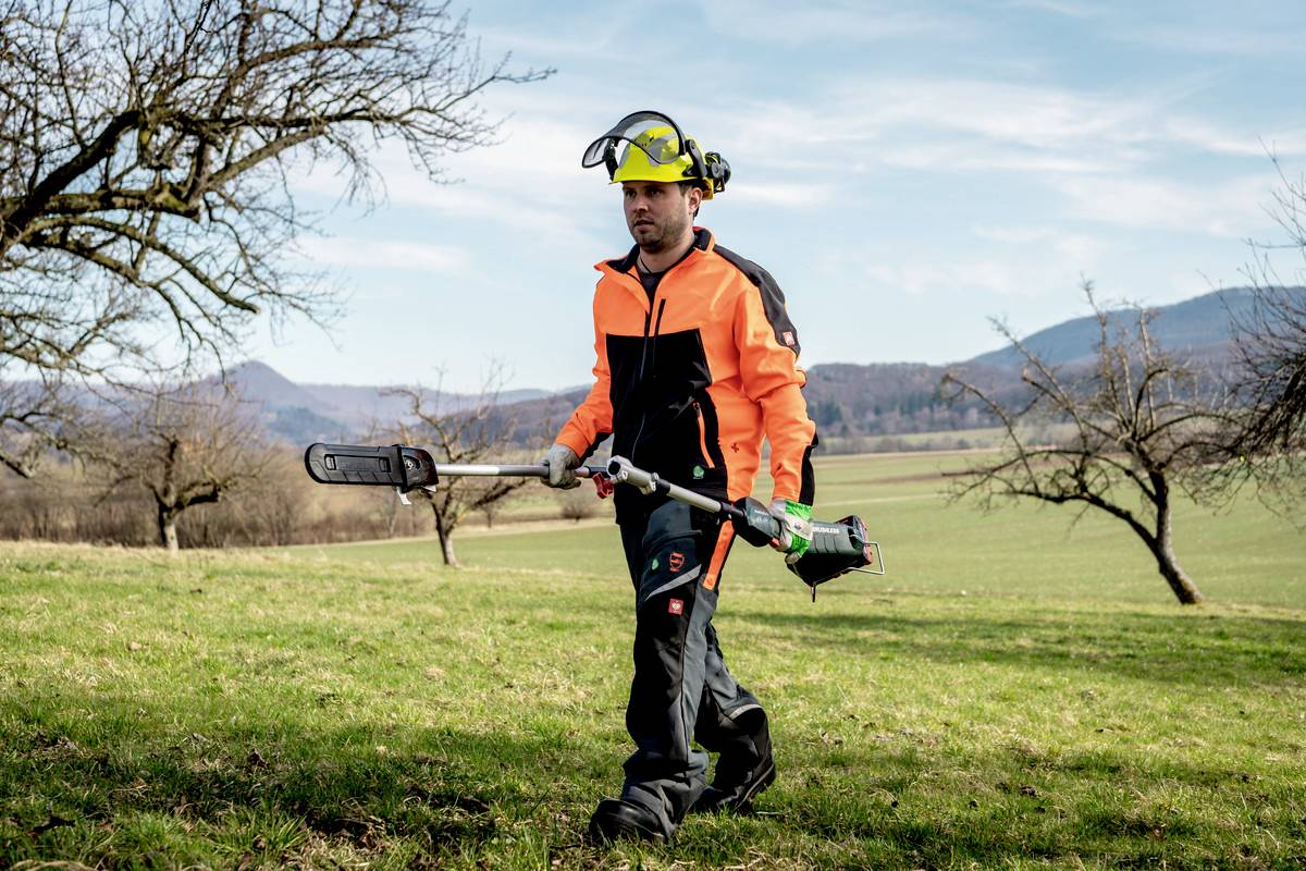 Ein Mann in Schutzkleidung mäht Gras auf einer Wiese mit einem Freischneider. Im Hintergrund sind kahle Bäume und Berge zu sehen.