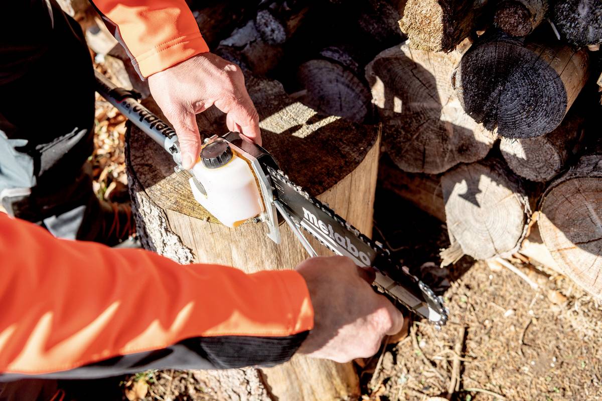 Eine Person schärft mit einer Feile die Kette einer Motorsäge, die auf einem Baumstumpf liegt. Im Hintergrund sind Holzscheite zu sehen.