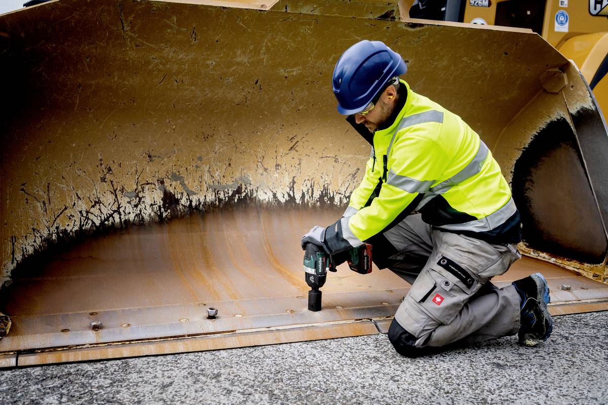 Ein Bauarbeiter in Sicherheitskleidung kniet und verwendet ein Elektrowerkzeug, um eine große Metalllöffelschaufel zu reparieren.