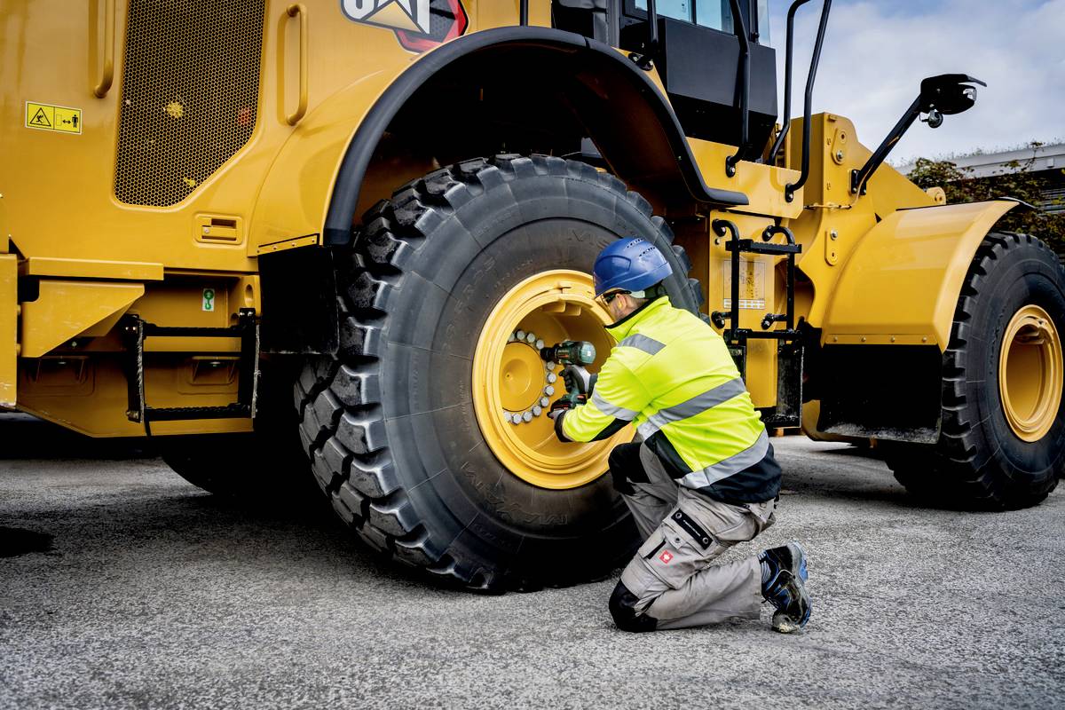 Eine Person in Schutzkleidung überprüft oder repariert den Reifen eines großen gelben Baustellenfahrzeugs auf einem Asphaltplatz.
