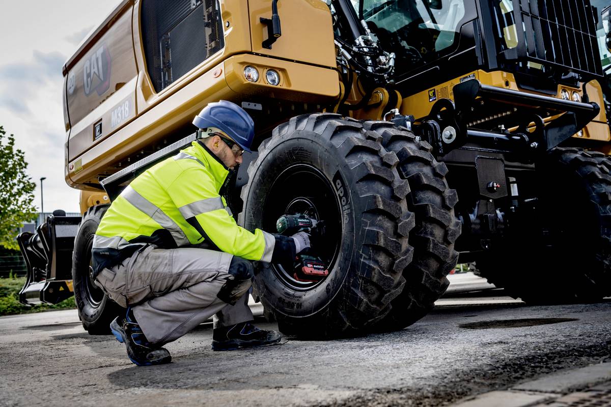 Ein Arbeiter in Schutzkleidung repariert ein großes Rad einer gelben Baumaschine. Die Umgebung deutet auf eine Baustelle hin.