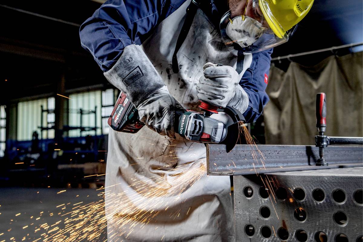 Eine Person in Schutzkleidung schleift mit einem Winkelschleifer Metall, Funken fliegen. Arbeitet in einer Werkstattumgebung.