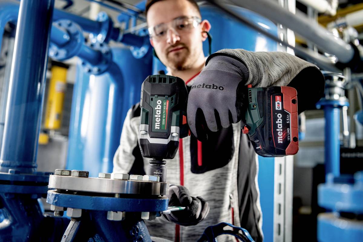 Ein Mann in Arbeitskleidung verwendet ein Metabo-Akkuwerkzeug, um eine Schraube an einer industriellen Maschine anzuziehen.