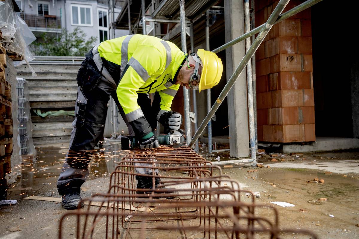 Bauarbeiter mit gelber Weste und Helm schneidet Metallstangen mit einem Winkelschleifer. Funken fliegen. Baustelle im Hintergrund.
