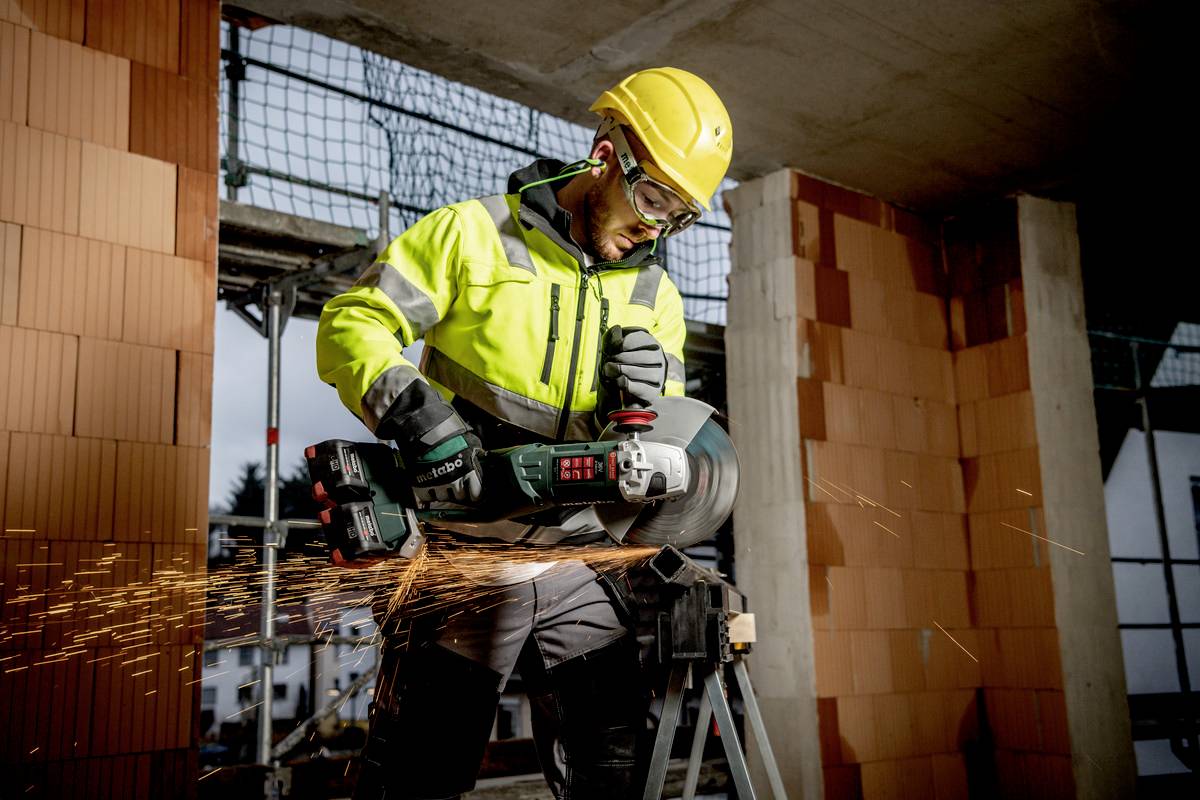 Ein Bauarbeiter mit Schutzkleidung schneidet mit einem Winkelschleifer Beton, Funken sprühen. Baustelle im Hintergrund.