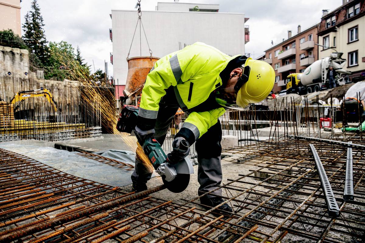 Ein Bauarbeiter in Schutzkleidung schneidet Stahlstäbe auf einer Baustelle. Im Hintergrund sind Gebäude und Baumaschinen sichtbar.