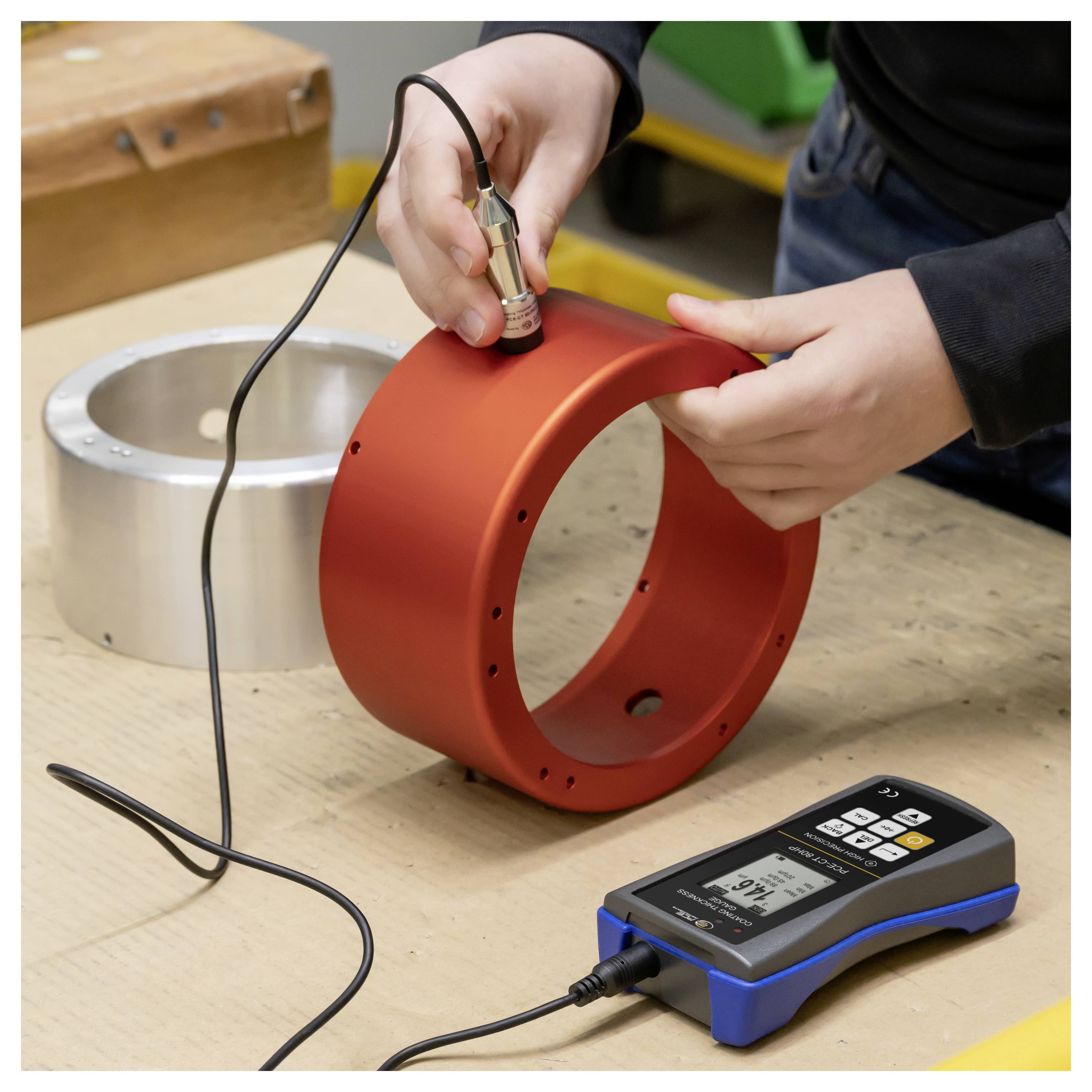 Person, die mit einer tragbaren Messsonde und einem digitalen Messinstrument die Oberfläche eines roten Metallrings auf einer Werkbank misst.