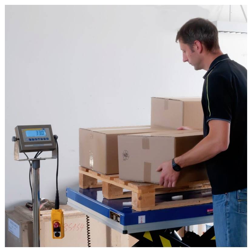 Mann in einem Lagerhaus positioniert Kartonboxen auf einer Palettenwaage, wobei eine digitale Gewichtsanzeige sichtbar ist.