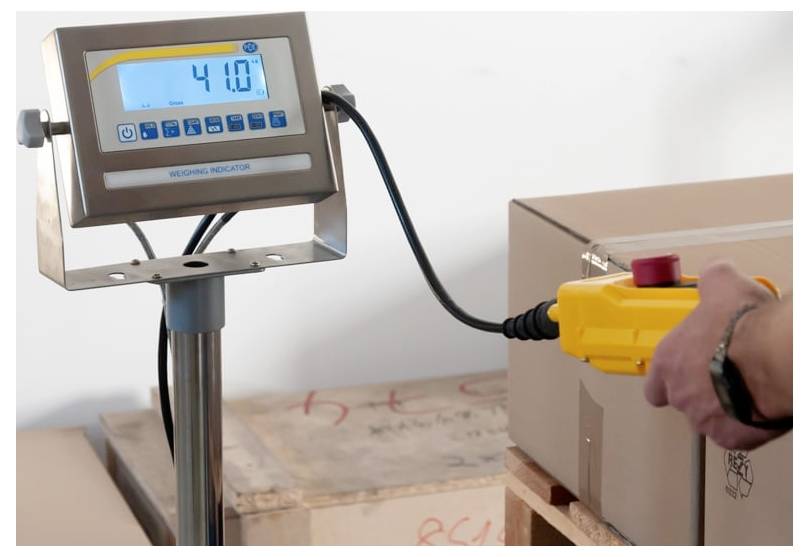 Digitale Wägeanzeige, die 4,10 anzeigt, über ein Kabel mit einem gelben Steuergerät neben einem verpackten Paket auf einer Palette verbunden.