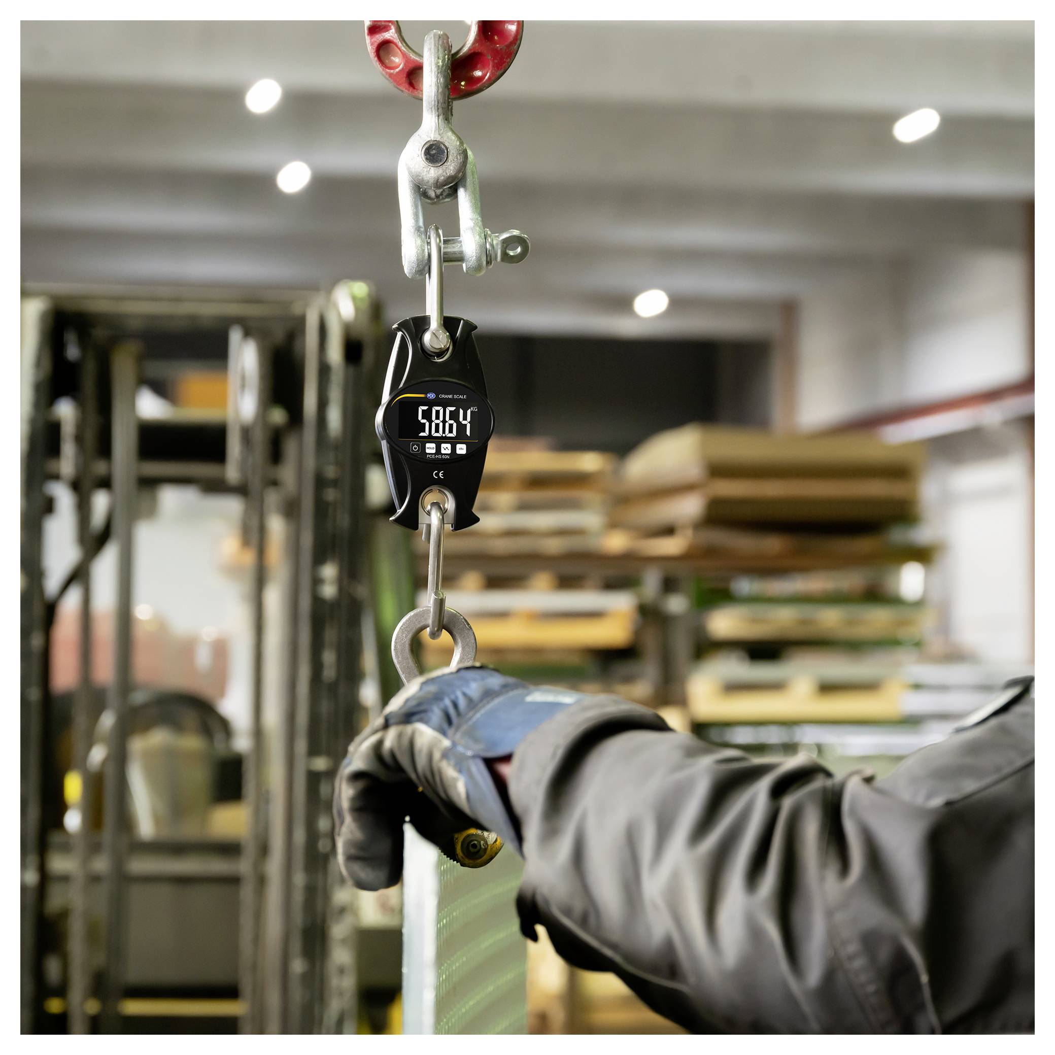 Handschuhbepackter Arbeiter, der eine hängende Last bearbeitet, die an einer digitalen Hängewaage befestigt ist und 5864 anzeigt, in einem Industrielager.
