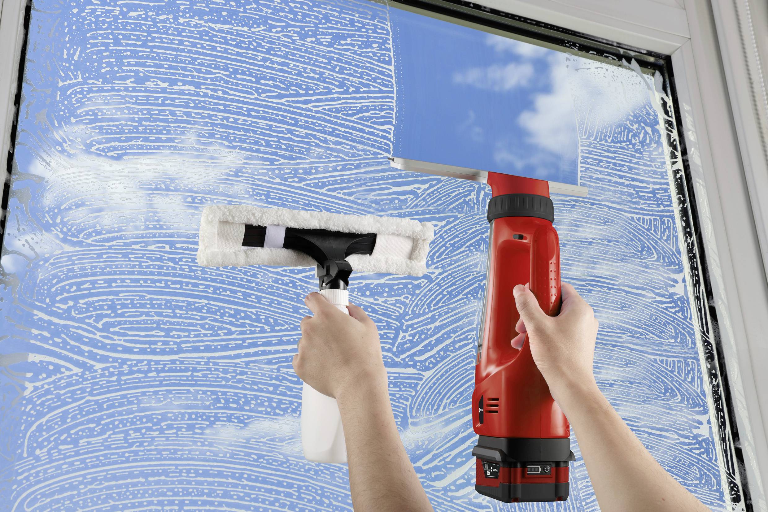 Eine Person reinigt ein Fenster mit einem Sprühreiniger und einem Fensterabzieher. Der Himmel spiegelt sich im Glas, Wolken sind sichtbar.