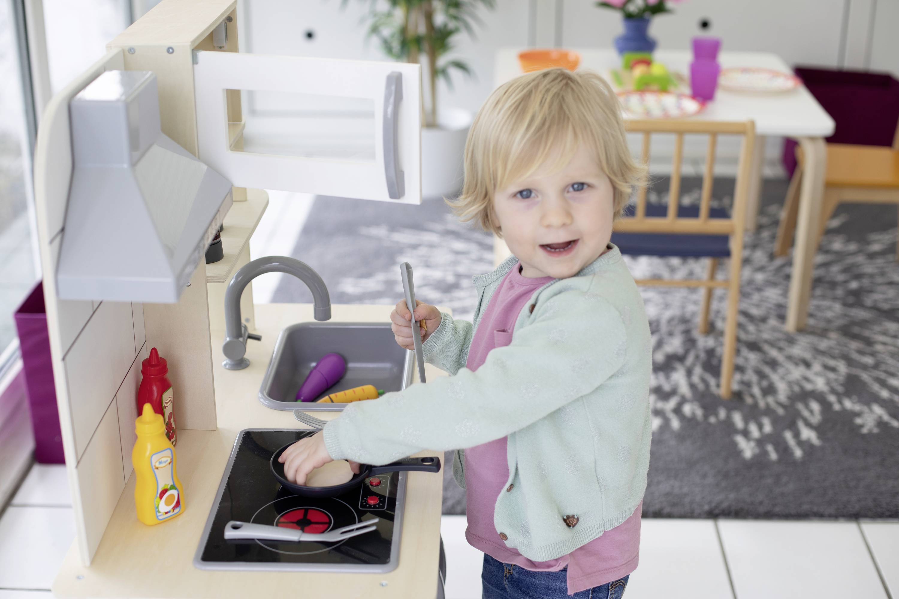 Ein Kind spielt in einer Spielküche und benutzt eine Pfanne auf dem Herd. Im Hintergrund ist ein Tisch mit Geschirr zu sehen.