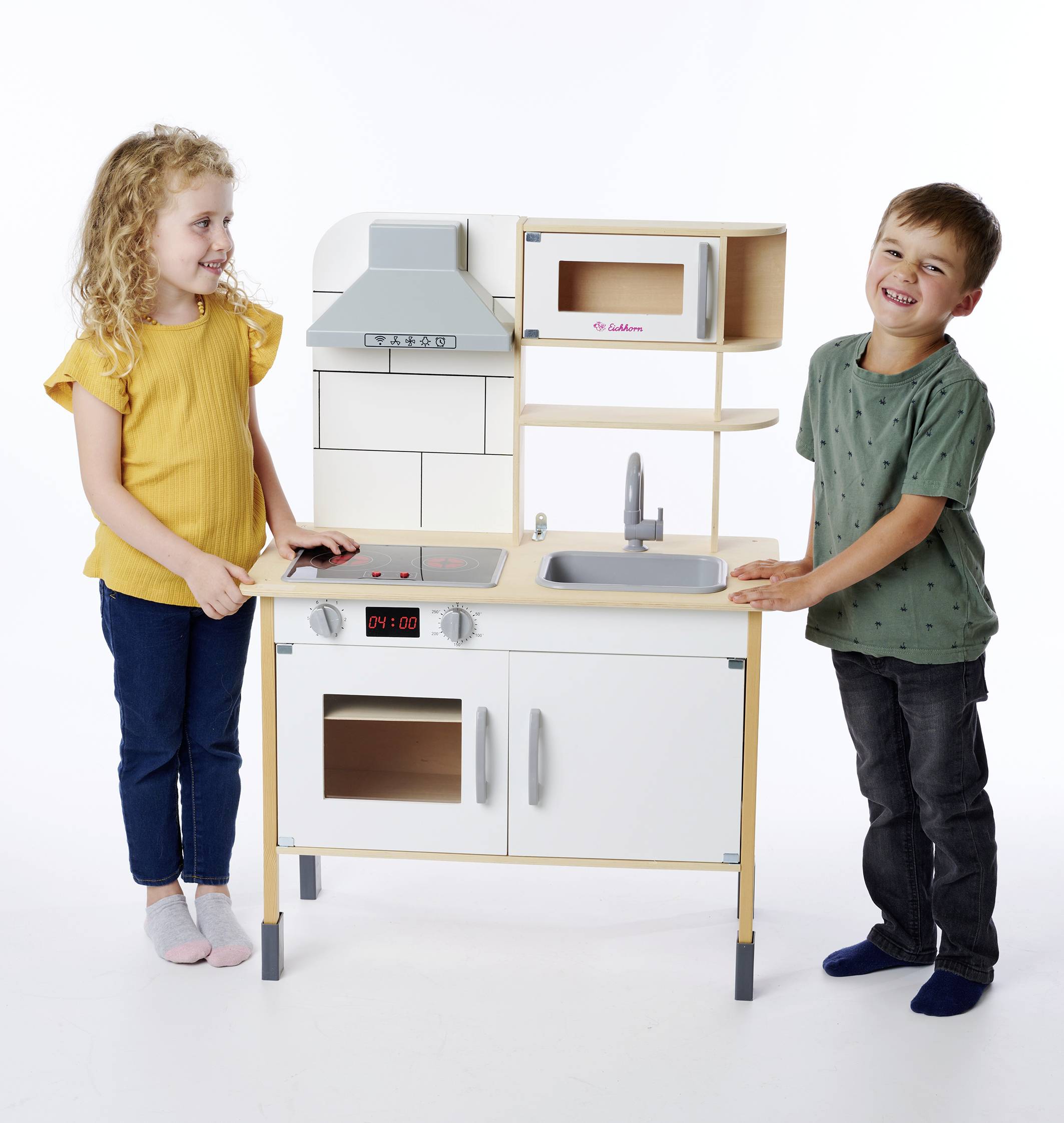 Zwei Kinder spielen glücklich mit einer Spielküche aus Holz, bestehend aus Herd, Spüle und Schränken. Mädchen trägt ein gelbes Shirt.