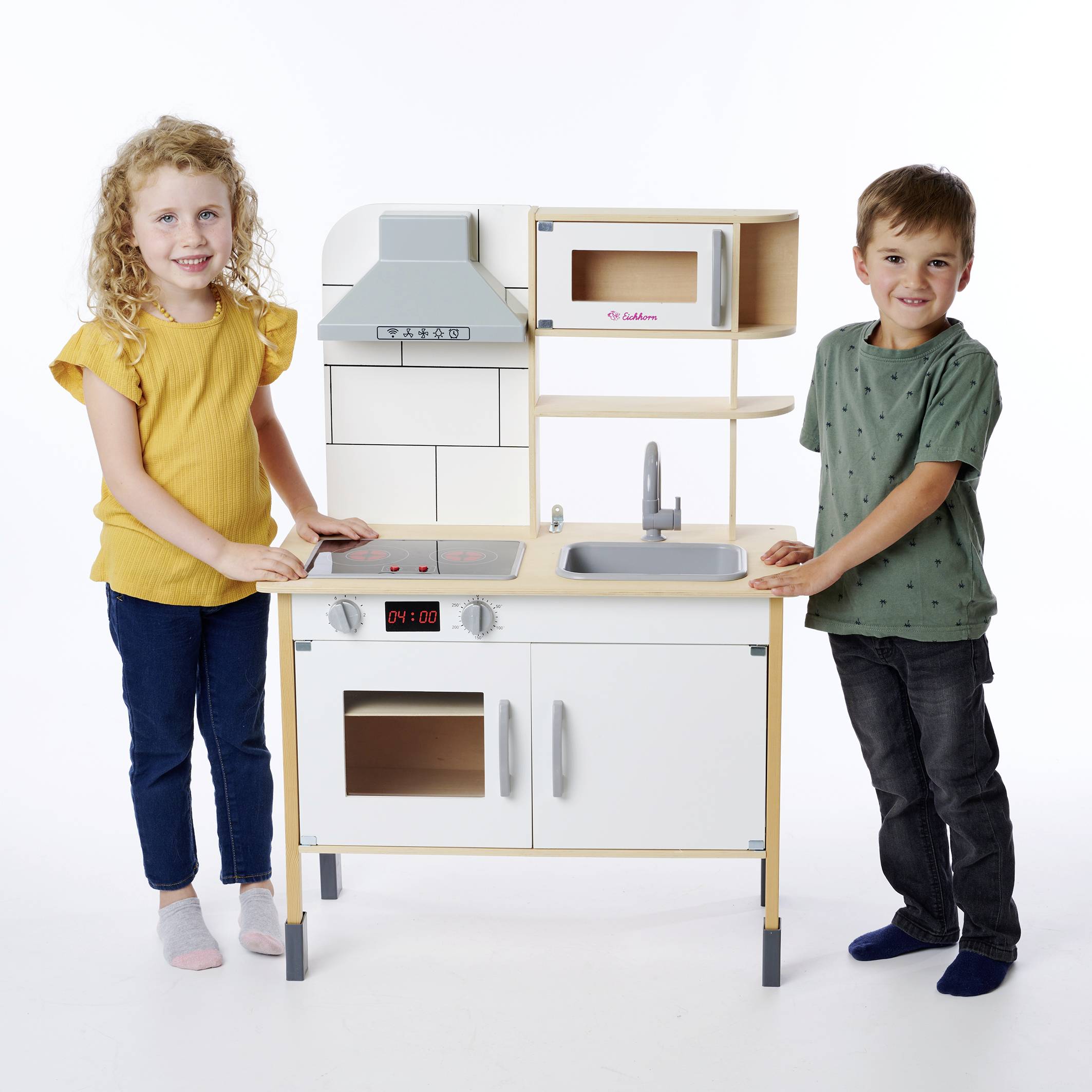 Ein Mädchen und ein Junge stehen neben einer Spielküche aus Holz. Das Mädchen trägt ein gelbes Oberteil, der Junge ein grün gepunktetes. Beide lächeln.