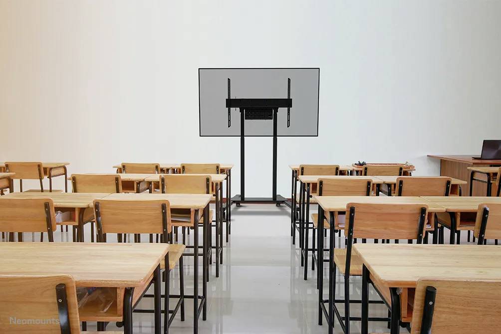 Klassenzimmer mit leeren Holztischen und -stühlen. Ein großer Bildschirm vorne. Links und rechts sind Wände und ein Lehrerpult zu sehen.