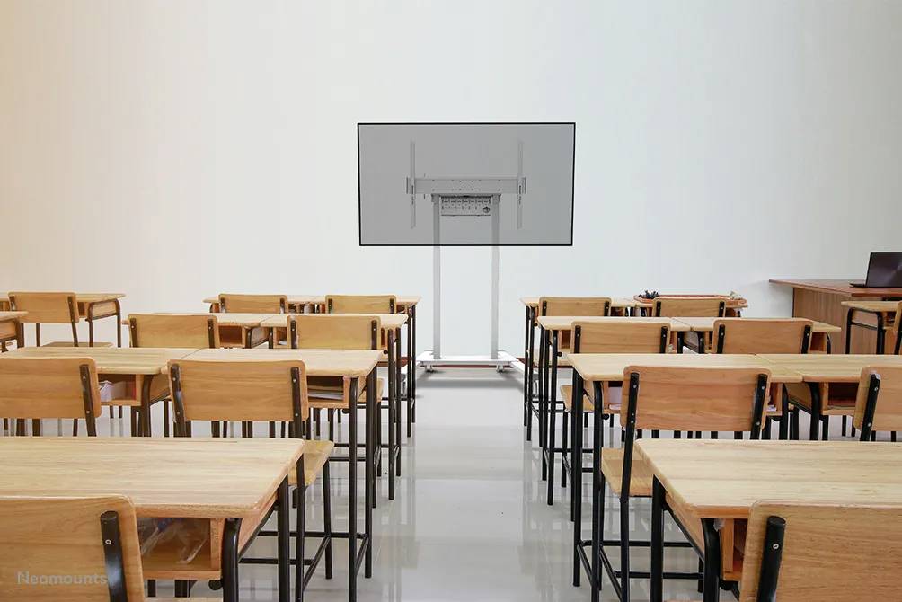 Klassenzimmer mit leeren Holzstühlen und Tischen, einem großen Display vorne und einem Lehrertisch rechts im Bild.