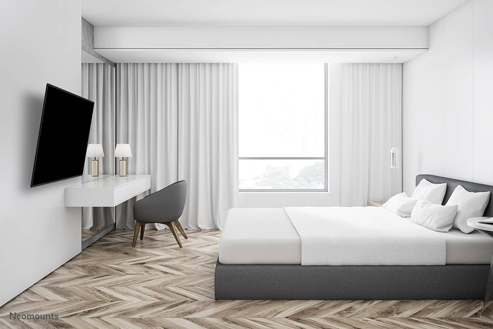 Helles Schlafzimmer mit großem Fenster, grauem Bett, Schreibtisch, Stuhl und Wandfernseher. Holzboden und weiße Vorhänge.