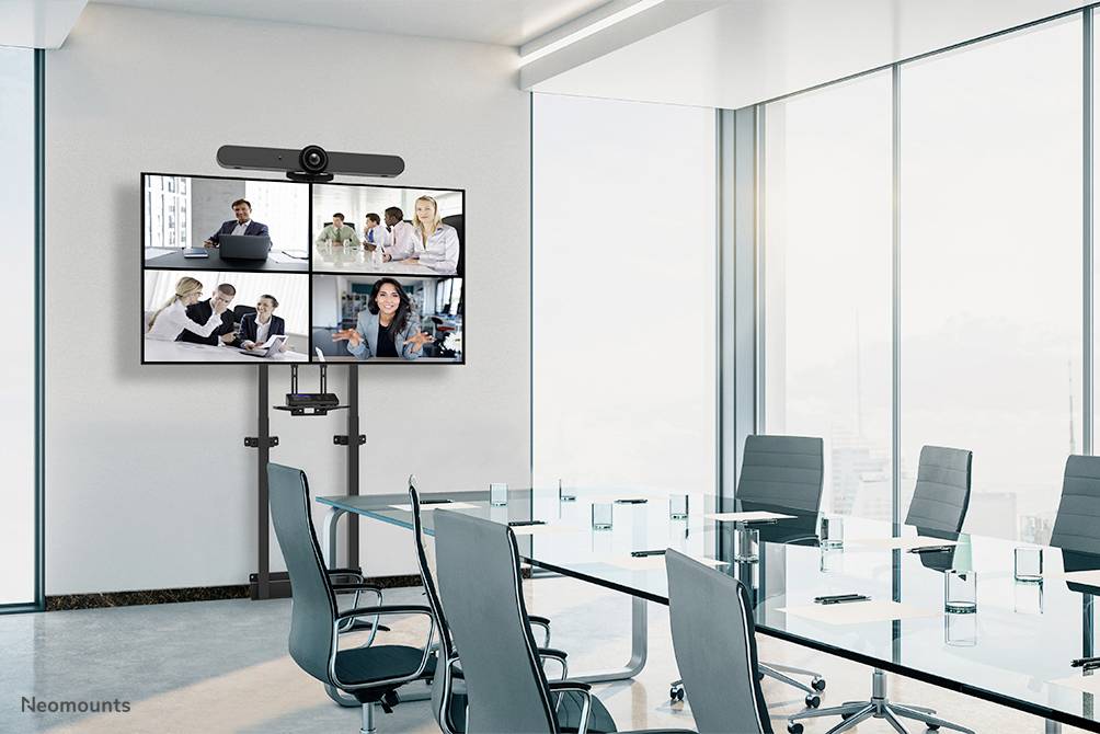 Konferenzraum mit großem Bildschirm zeigt Videokonferenz mit vier Teilnehmergruppen. Moderner Tisch und Stühle im Vordergrund.