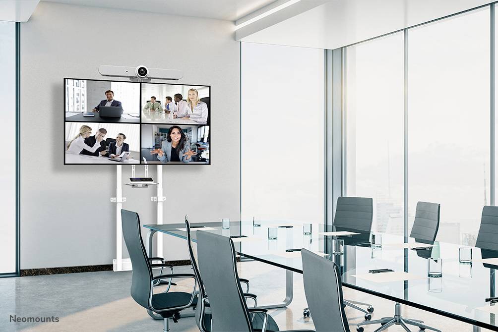 Konferenzraum mit einem großen Bildschirm, der vier Videokonferenz-Teilnehmer zeigt. Moderne Möbel und leere Stühle sind sichtbar.