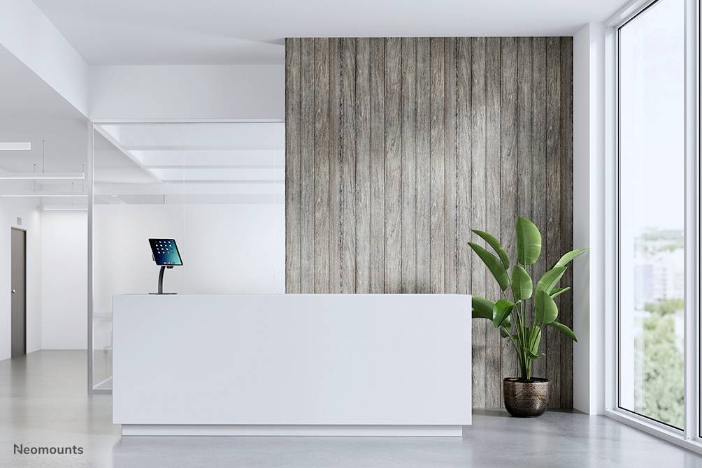 Großes Büro mit weißem Empfangstresen, Tablet auf Ständer, große Fenster rechts, Holzverkleidung an der Wand und Pflanze im Topf links.