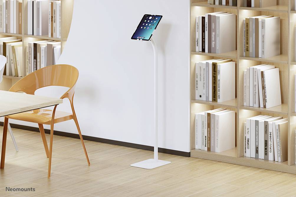 Ein Tablet steht auf einem weißen Ständer neben einem leeren orangefarbenen Stuhl vor Regalen mit Büchern in einem modernen Raum.