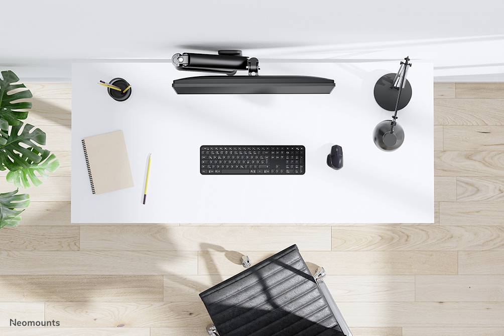 Ein aufgeräumter Schreibtisch mit Tastatur, Maus, Lampe und Notizbuch, daneben eine Pflanze und ein schwarzer Bürostuhl.