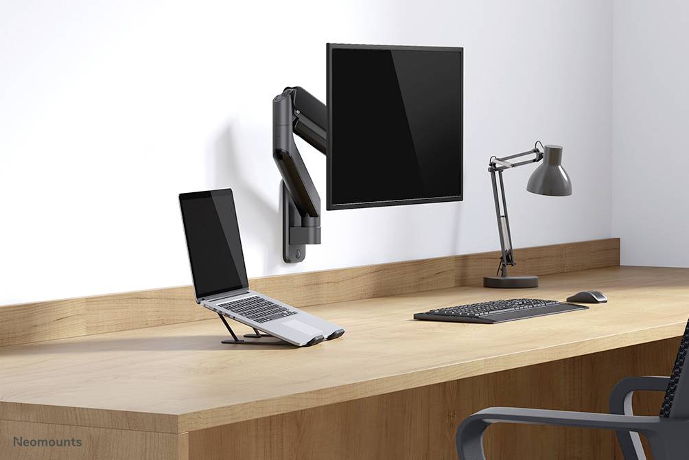 Ein aufgeräumter Schreibtisch mit Laptop auf einem Ständer, einem festmontierten Monitor an der Wand, einer Tastatur und einer Schreibtischlampe.
