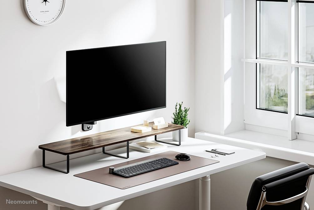 Ein minimalistischer Schreibtisch mit Monitor auf Holzständer, Tastatur, Maus, Dokumenten und kleiner Pflanze, direkt neben einem Fenster.