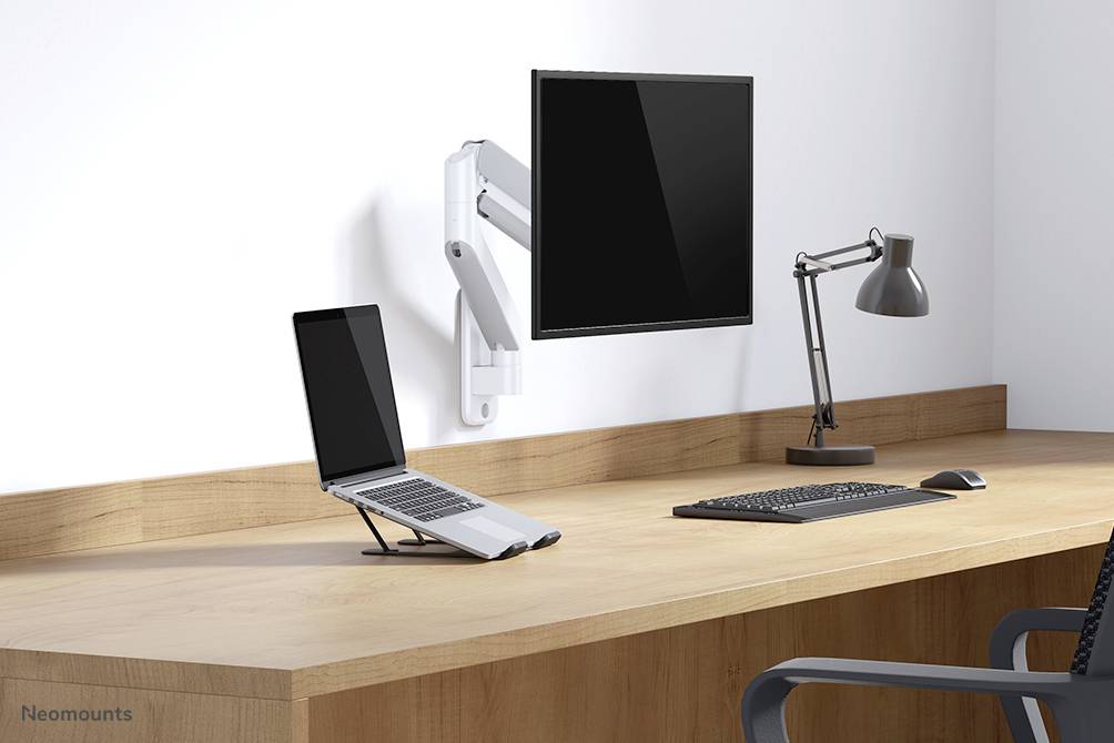 Ein Schreibtisch mit einem an der Wand montierten Monitor, einem Laptop auf einem Ständer, einer Tastatur und einer Lampe.