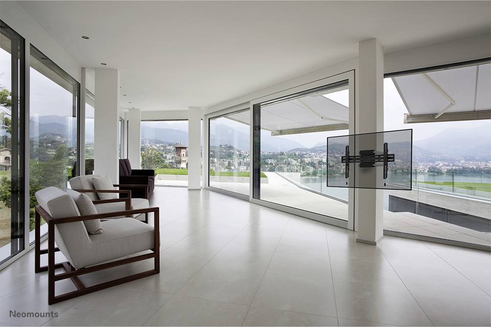 Ein heller, moderner Raum mit großen Fenstern bietet Blick auf eine malerische Landschaft. Zwei weiße Sessel stehen links im Raum.