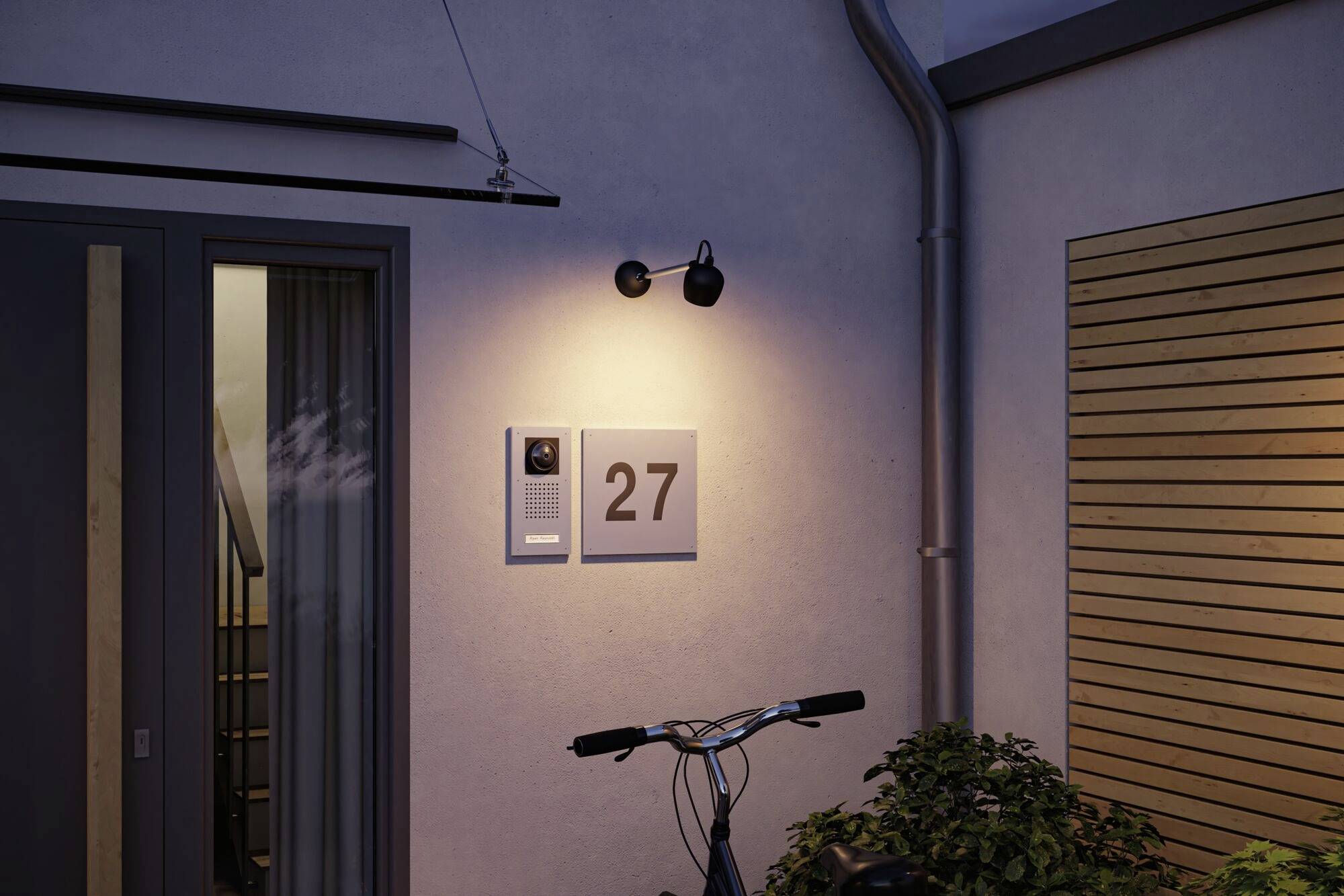 Wohnhaus bei Dämmerung, Ansicht eines Eingangs mit der Nummer '27', Gegensprechanlage und Kamera; Fahrrad und Pflanzen im Vordergrund.