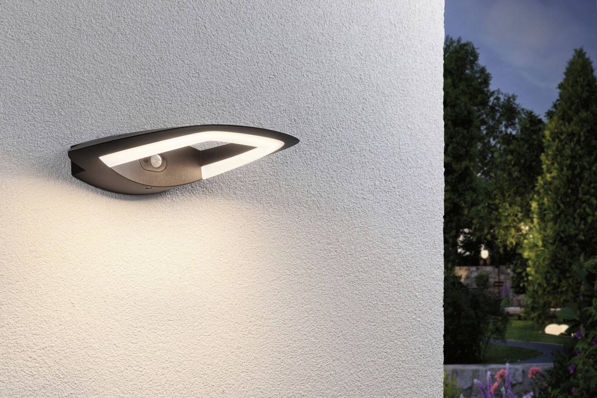 Außenwandleuchte mit modernem Design beleuchtet eine weiße Wand, umgeben von einem Garten in der Abenddämmerung.