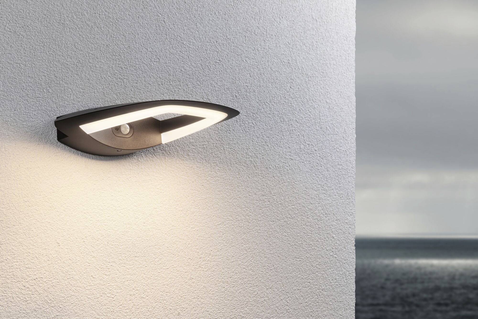 Eine moderne Außenleuchte an einer weißen Wand beleuchtet dezent die Umgebung. Im Hintergrund ist das Meer zu sehen.