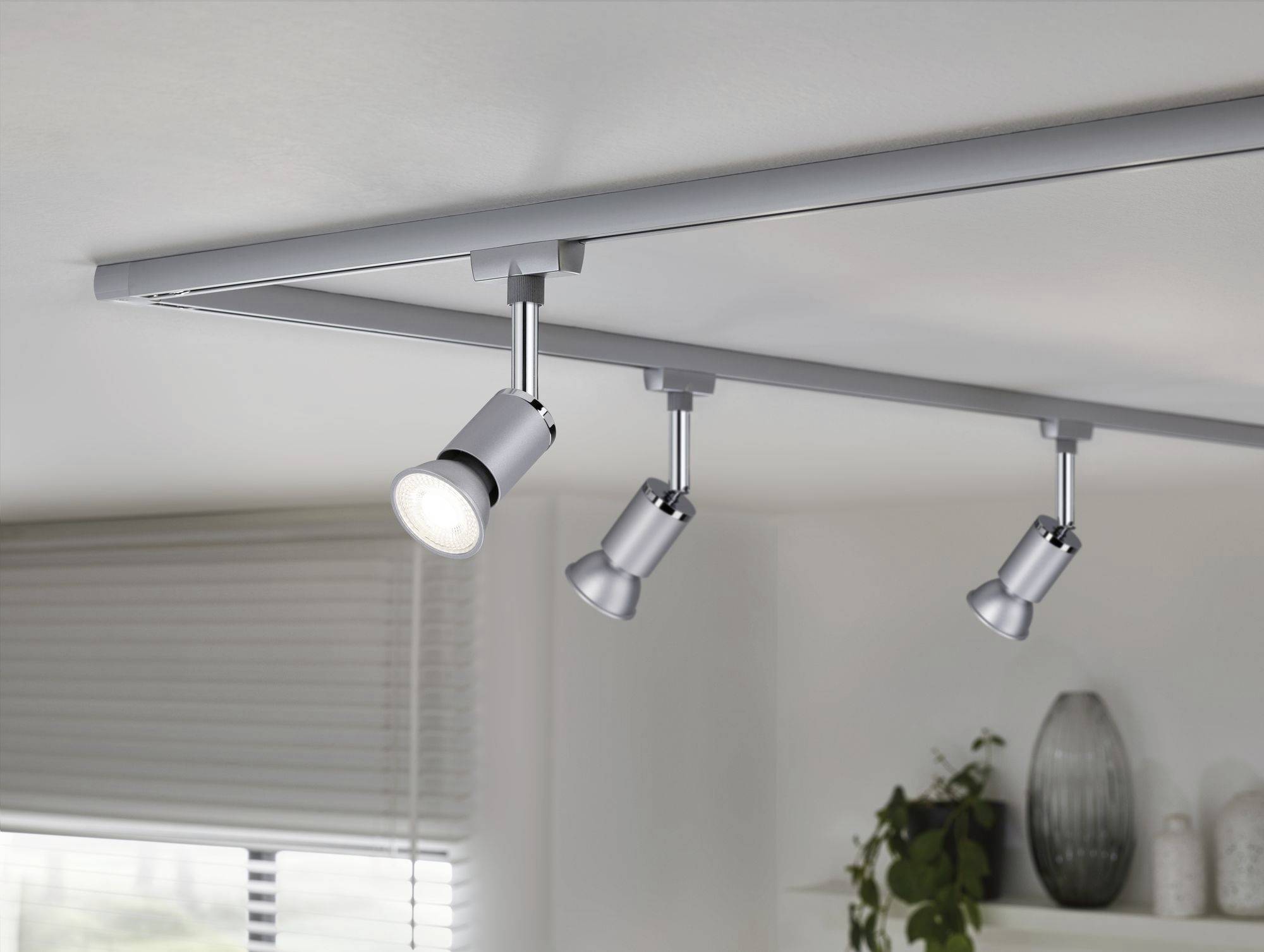Eine moderne Deckenleuchte mit drei verstellbaren Spotlights, montiert in einem Wohnzimmer. Im Hintergrund sind Pflanzen und Keramikvasen.