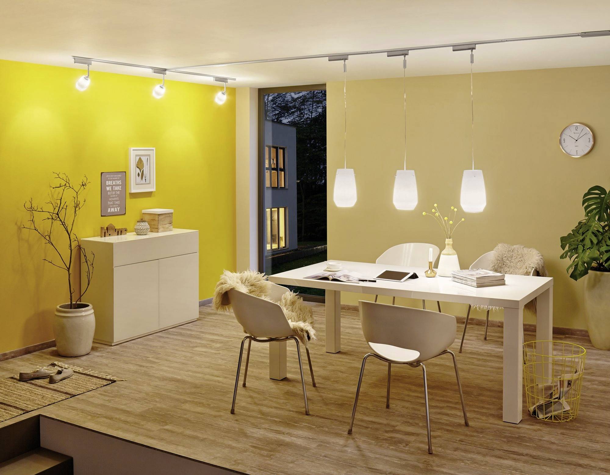 Esszimmer mit modernem Interieur, gelb gestrichene Wände, weißer Tisch, vier Stühle, Pflanzen und Wanduhren. Fenster zeigt Abendhimmel.
