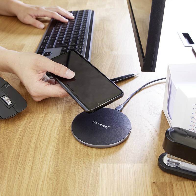 Eine Person lädt ein Smartphone kabellos auf einer runden Ladestation auf. Neben dem Smartphone liegt eine Computermaus und eine Tastatur.