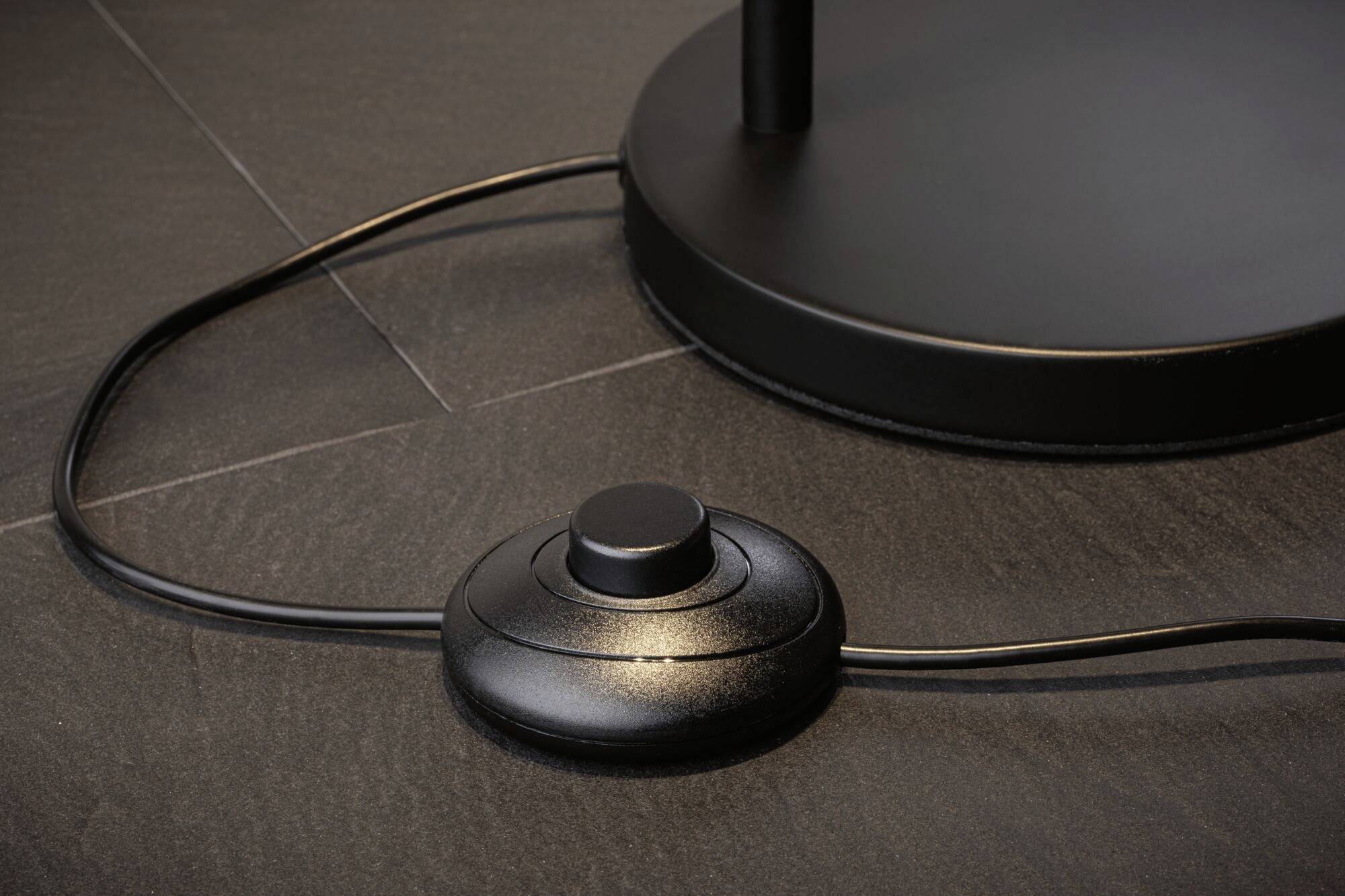 Schwarzer Fußschalter auf einem dunklen Boden, mit Kabel verbunden. Der Schalter steuert ein elektrisches Gerät.