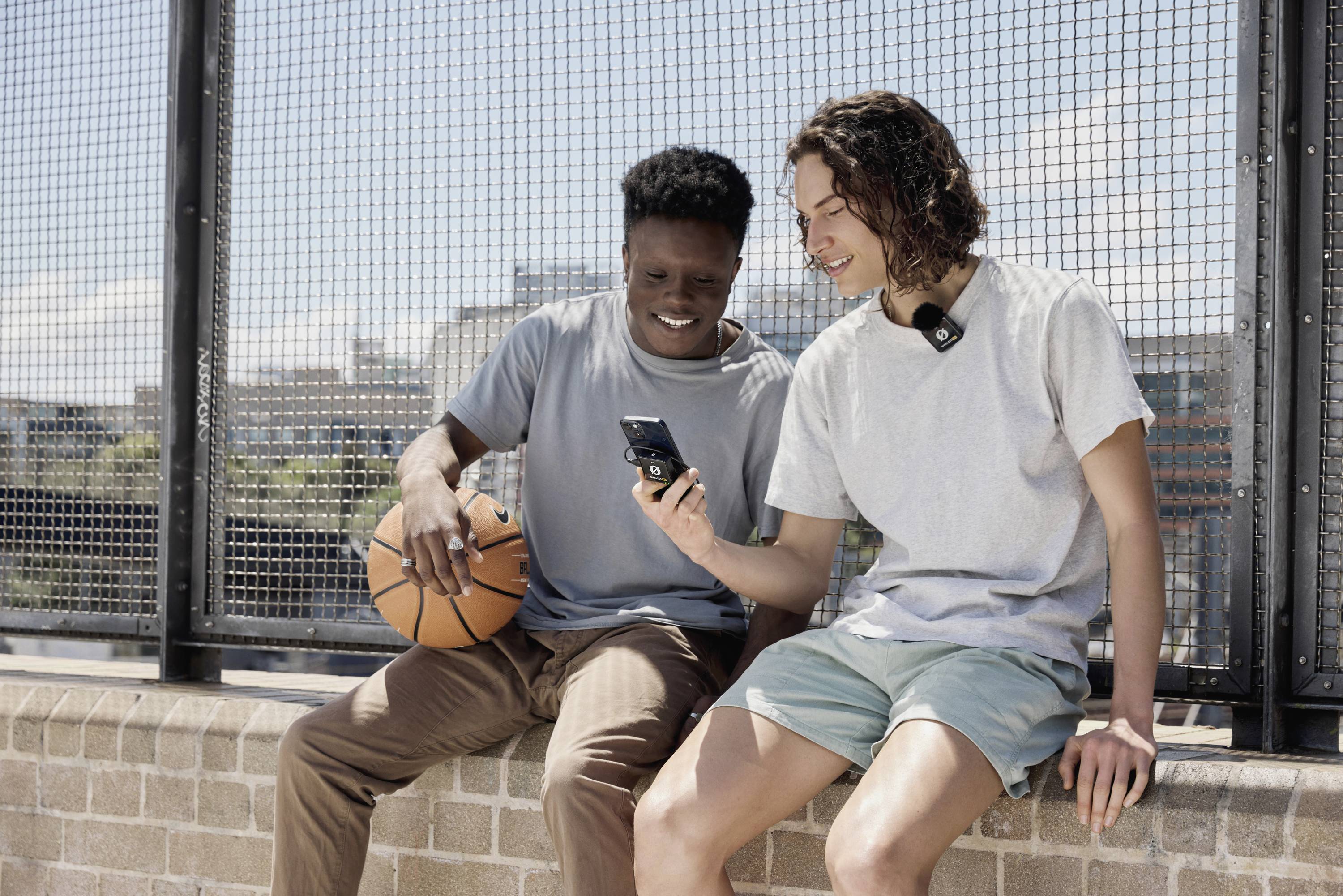Zwei junge Männer sitzen auf einer Mauer. Einer hält einen Basketball, der andere zeigt ihm etwas auf einem Smartphone. Sie wirken entspannt.