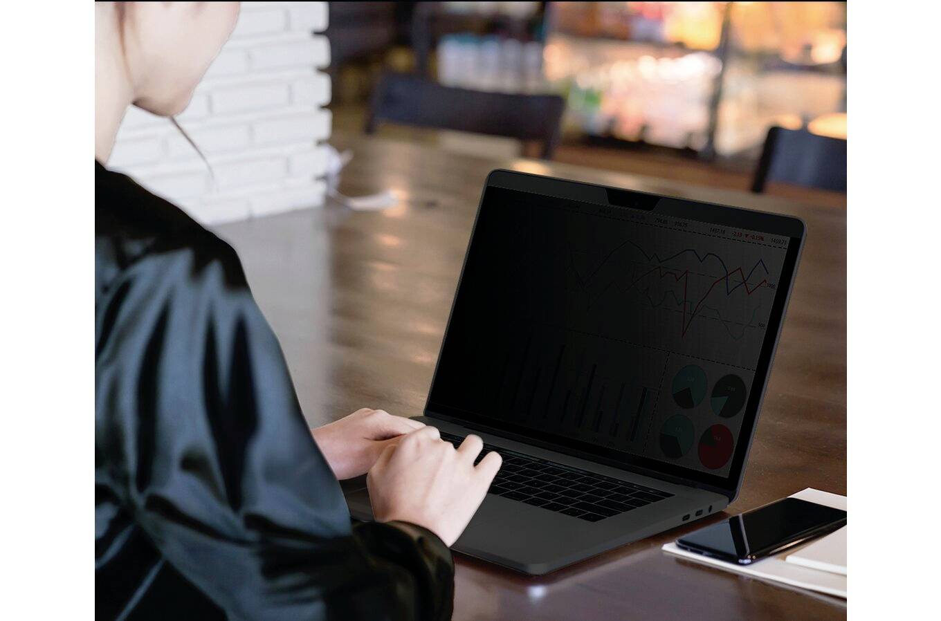 Person arbeitet an einem Laptop in einem Café. Auf dem Bildschirm sind Diagramme und Grafiken zu sehen. Ein Smartphone liegt daneben.
