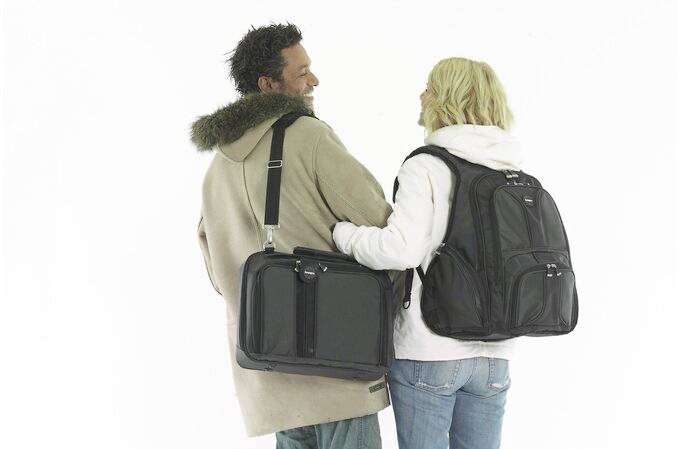 Eine Frau und ein Mann stehen lächelnd Rücken an Rücken. Die Frau trägt einen großen schwarzen Rucksack und der Mann eine schwarze Umhängetasche.