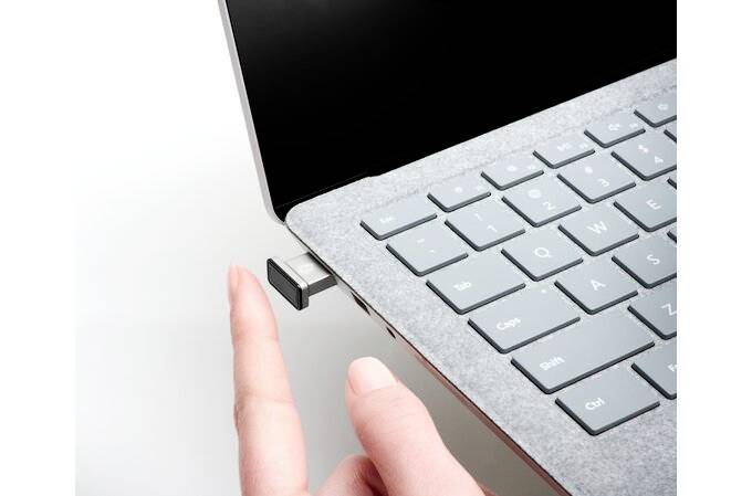 Eine Hand zeigt auf ein USB-Gerät, das an einen Laptop angeschlossen ist. Der Laptop ist von der Seite sichtbar und zeigt die Tastatur.