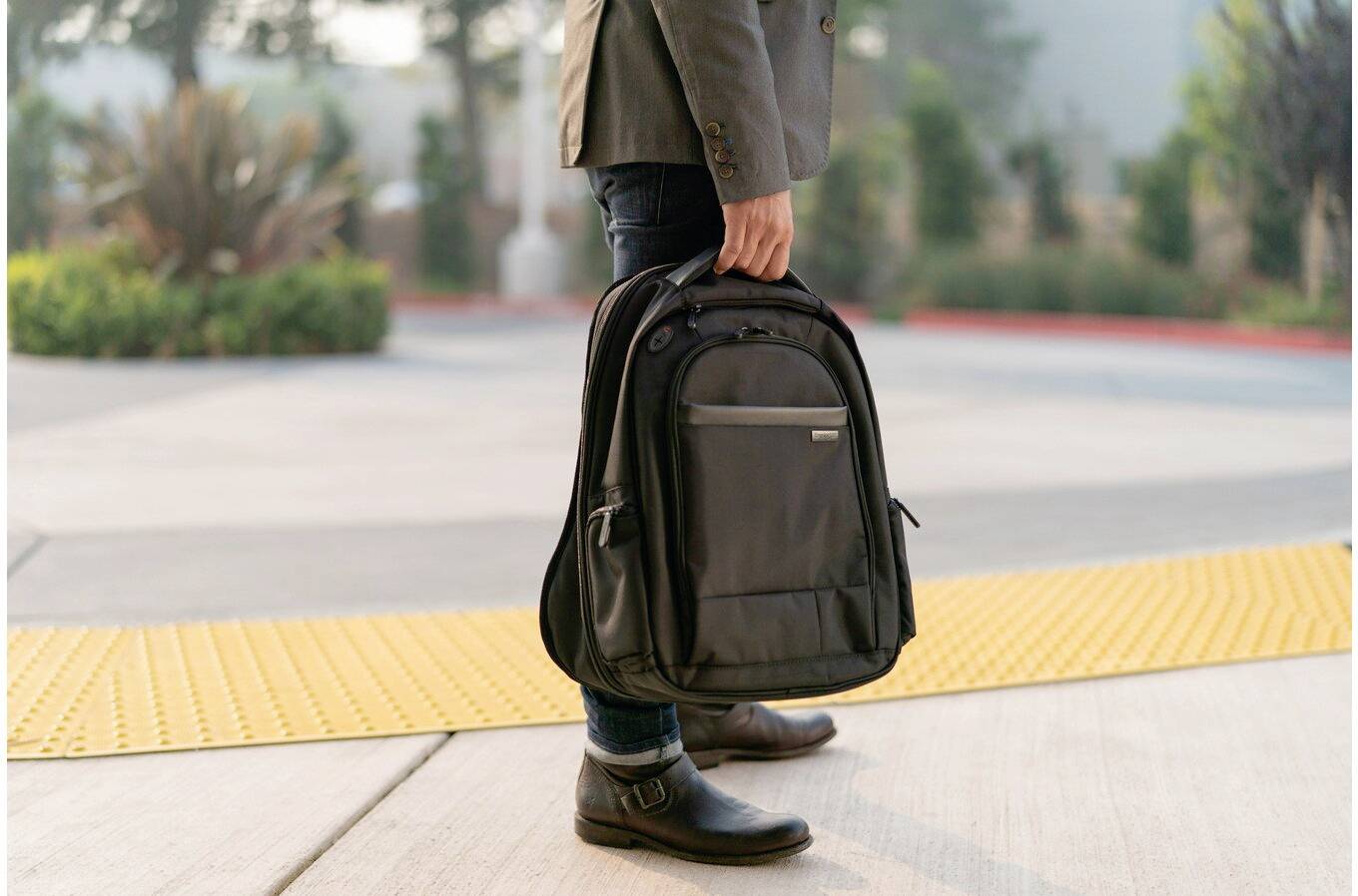 Eine Person geht über einen Bürgersteig und trägt einen schwarzen Rucksack in der Hand. Der Hintergrund ist leicht unscharf.