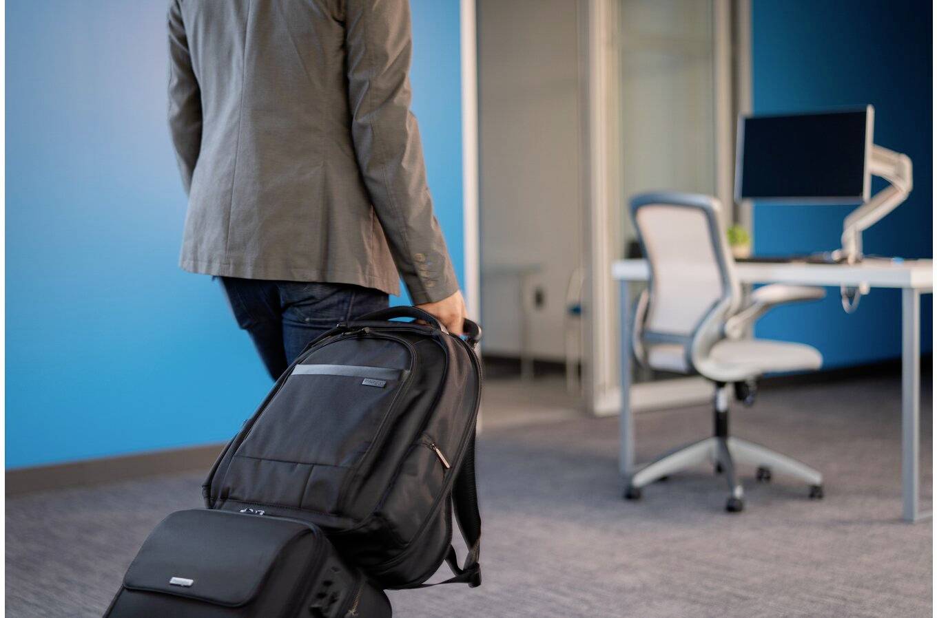 Eine Person zieht eine Rolltasche durch ein modernes Büro mit blau gestrichenen Wänden, einem Schreibtisch und einem Computer.