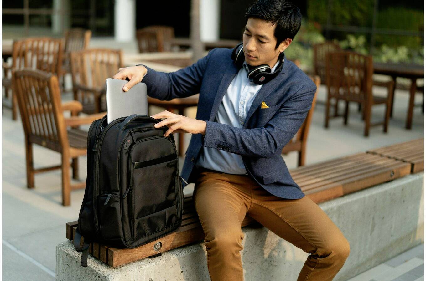 Ein Mann setzt sich auf eine Bank im Freien und steckt einen Laptop in seinen Rucksack. Er trägt Kopfhörer um den Hals.