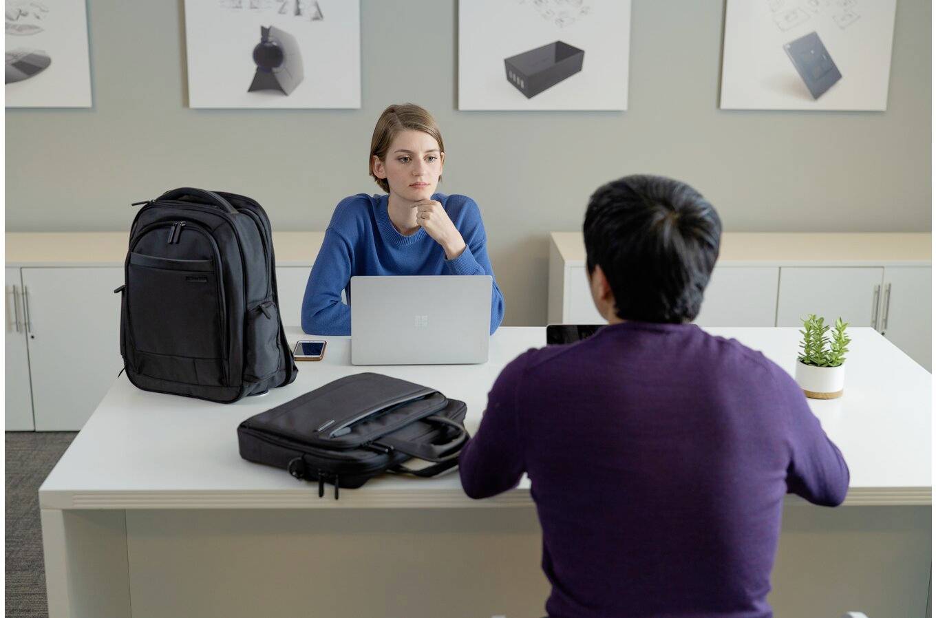 Zwei Personen sitzen an einem Tisch in einem Büroraum. Eine Person arbeitet am Laptop, während die andere zuhört. Auf dem Tisch liegen Taschen.