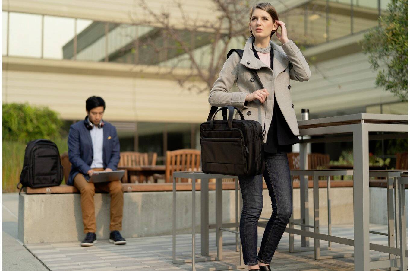 Eine Frau steht auf einer Terrasse, hält eine Aktentasche und telefoniert. Im Hintergrund sitzt ein Mann auf einer Bank mit einem Laptop.